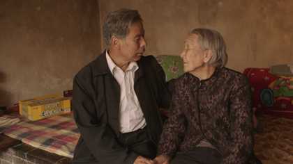 An older couple having a sweet conversation while sitting on a bed and holding hands.