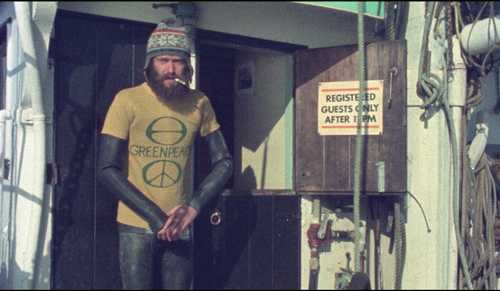 A man in a GreenPeace t-shirt and toque standing in the cold and smoking a cigarette.