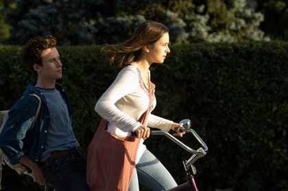 A teenage girl pedaling a bicycle with determination toward the setting sun while a teenage boy sits behind her.
